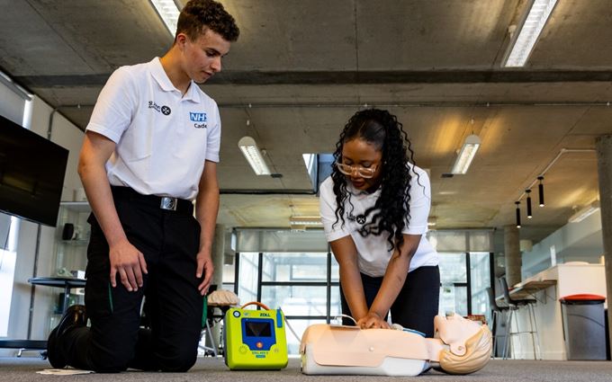 Two teenagers wearing an NHS Cadet polo shirt kneeling on the floor. One of them doing chest compressions on a mannequin with a defibrillator attached. Two teenagers wearing an NHS Cadet polo shirt kneeling on the floor. One of them doing chest compressions on a mannequin with a defibrillator attached.