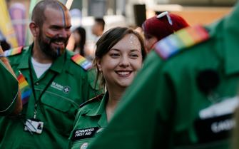 A smiling Advanced First Aider volunteer with glitter on their face and surrounded by other volunteers with rainbow epaulettes.  A smiling Advanced First Aider volunteer with glitter on their face and surrounded by other volunteers with rainbow epaulettes.
