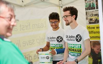 Two fundraising volunteers stood in a tent smiling. One is holding a collection bucket and the other is holding flyers. They're both facing a first aid volunteer. Two fundraising volunteers stood in a tent smiling. One is holding a collection bucket and the other is holding flyers. They're both facing a first aid volunteer.