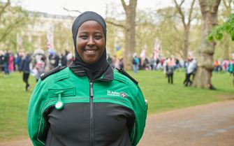 A volunteer standing in a busy park with their arms behind their back and smiling. A volunteer standing in a busy park with their arms behind their back and smiling.