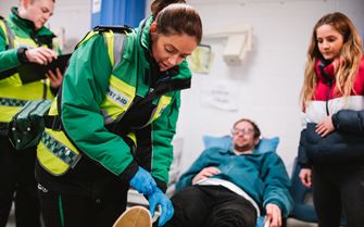 A patient is lying down on a bed being treated by a first aid volunteer. Another volunteer is stood looking at a folder.  A patient is lying down on a bed being treated by a first aid volunteer. Another volunteer is stood looking at a folder.